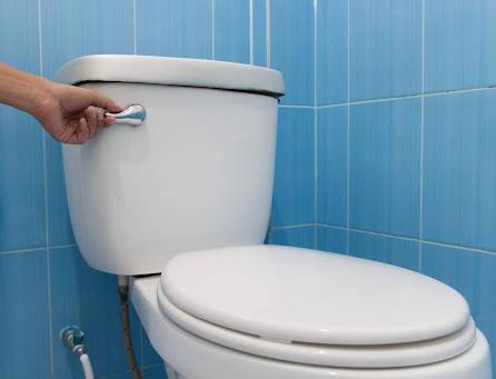 %Focuskw% | Strongbuilt Plumbing, Air, Electrical A hand is pressing the flush lever on a white toilet with a closed lid, set against blue tiled walls.