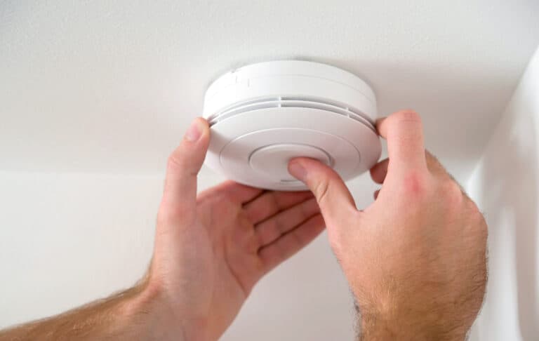 Hands installing a white smoke detector on a ceiling.