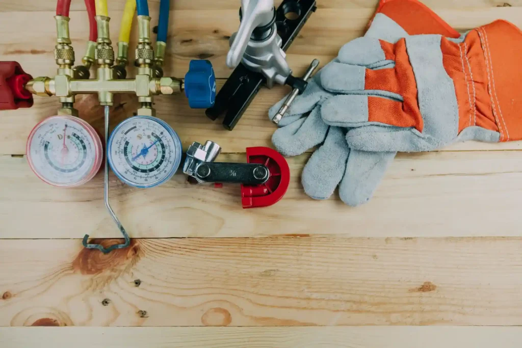 HVAC gauges, pipe cutter, valve, screwdriver, and a pair of protective gloves arranged on a wooden surface.