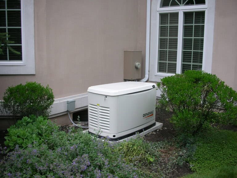 A standby generator installed outside a house, surrounded by bushes and plants, near a window with white blinds.