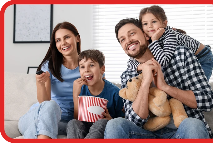 Family sitting on a couch watching TV. A woman holds a remote, a boy eats popcorn, a man smiles holding a girl and a teddy bear. They're all casually dressed and appear happy.