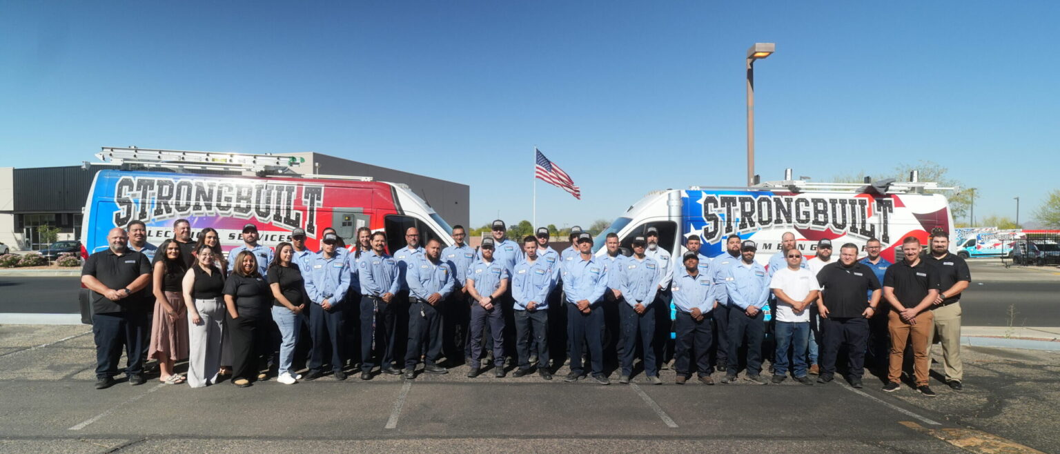 A group of people stand in front of two vans with "Strongbuilt" branding, parked in a lot with an American flag and buildings in the background.