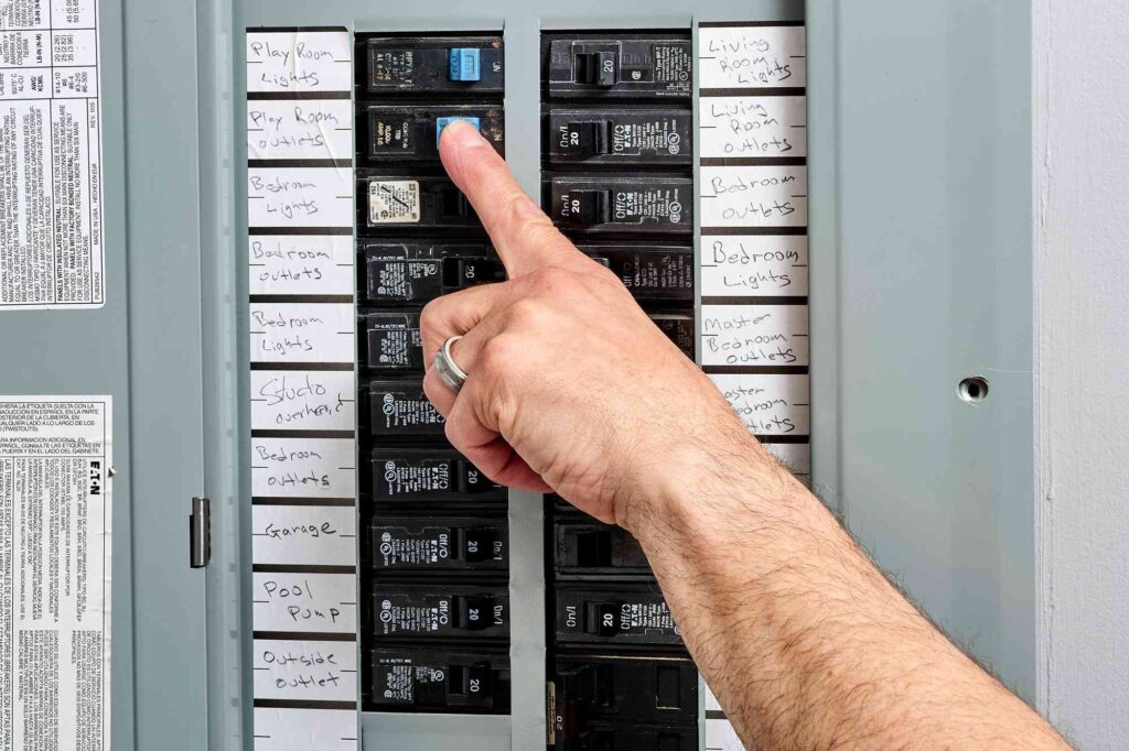 %Focuskw% | Strongbuilt Plumbing, Air, Electrical A hand with a ring points to a labeled circuit breaker in an electrical panel, with various switches labeled for different rooms and outlets.