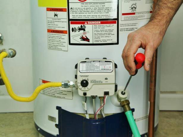A person uses a screwdriver to adjust a valve on a water heater, with warning labels and pipes visible.