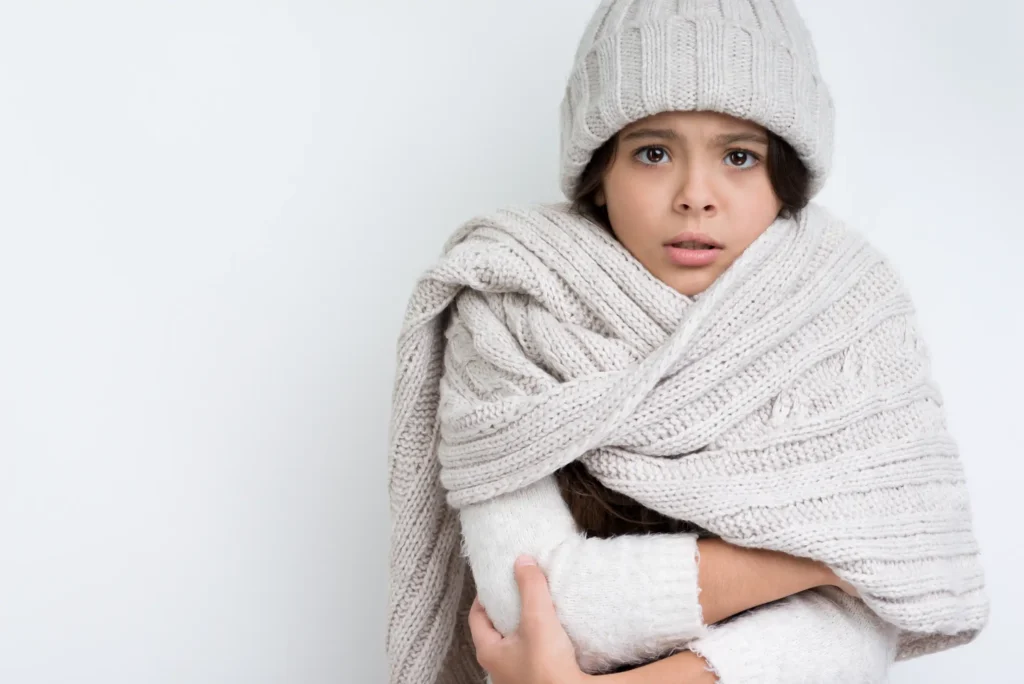 A young girl in a knit hat and large scarf hugs herself and looks cold against a plain white background.