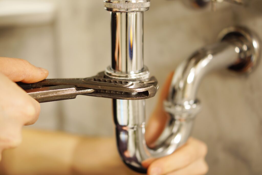 %Imagetitle% | Strongbuilt Plumbing &Amp; Air A person uses a wrench to tighten a metal pipe under a sink, focusing on plumbing maintenance.