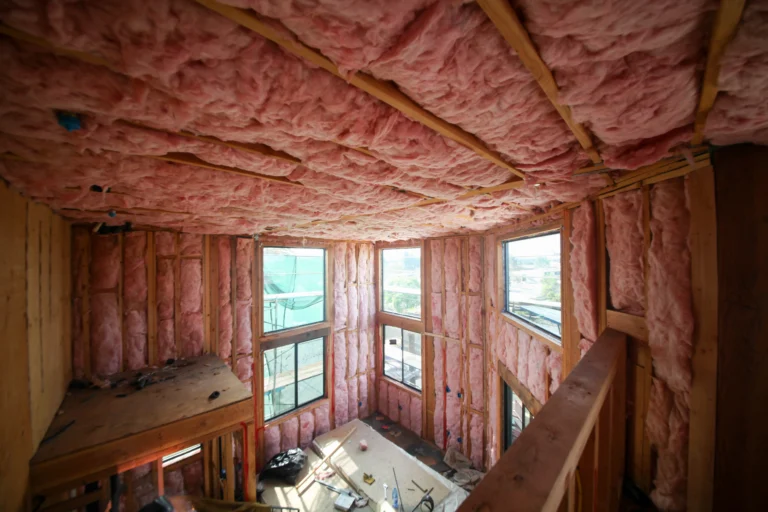 Interior of a house under construction with pink fiberglass insulation installed in the walls and ceiling, unfinished wood framing, visible heating installation components, and construction materials scattered on the floor.